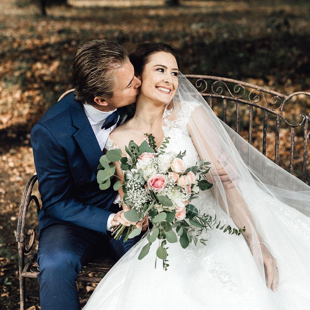 Eine Braut und ein Bräutigam teilen sich einen innigen Kuss auf einer rustikalen Bank inmitten des bezaubernden Waldes.