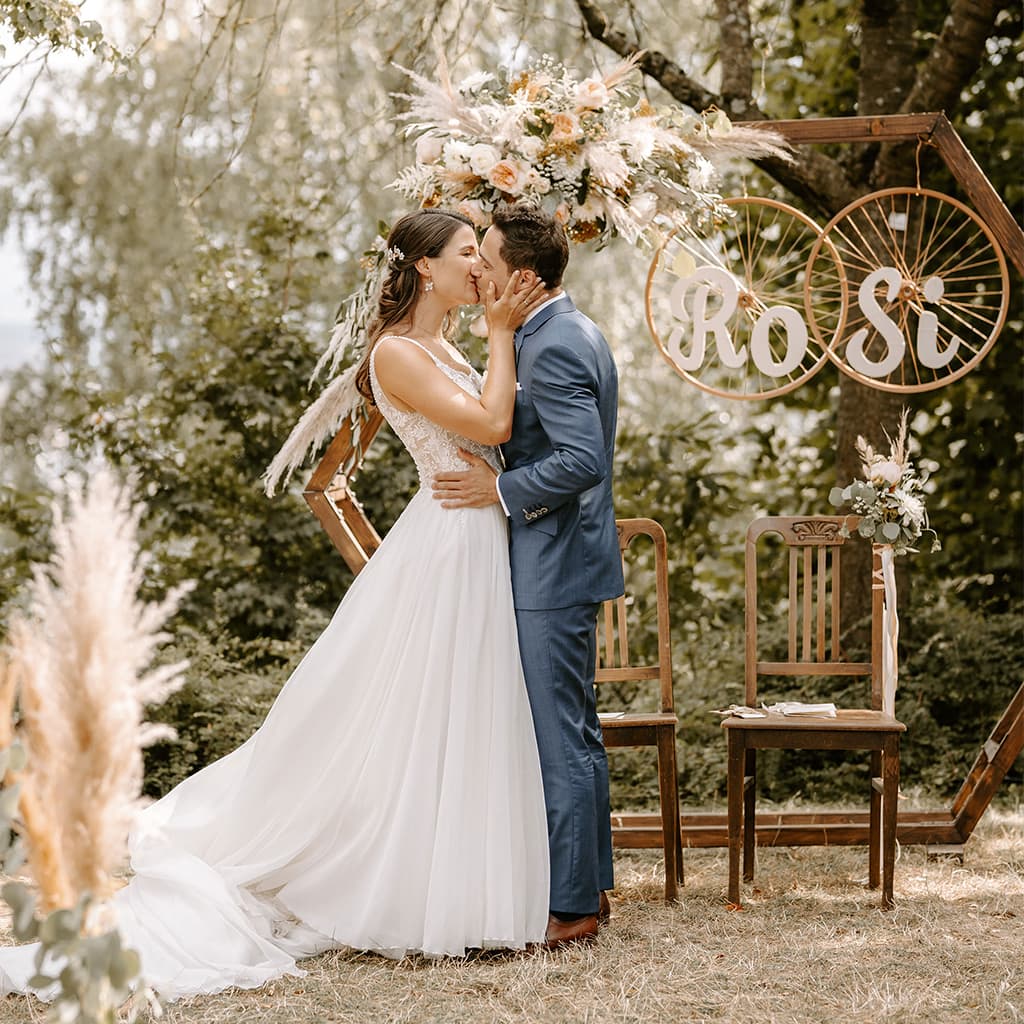Eine Braut und ein Bräutigam küssen sich vor einem hölzernen Hochzeitsbogen.