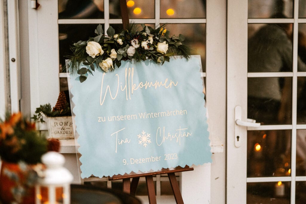 Ein Willkommensschild für eine winterliche Veranstaltung mit Blumenschmuck und dem Text „Willkommen zu unserem Wintermärchen – tina & christian – 9. Dezember 2023“.
