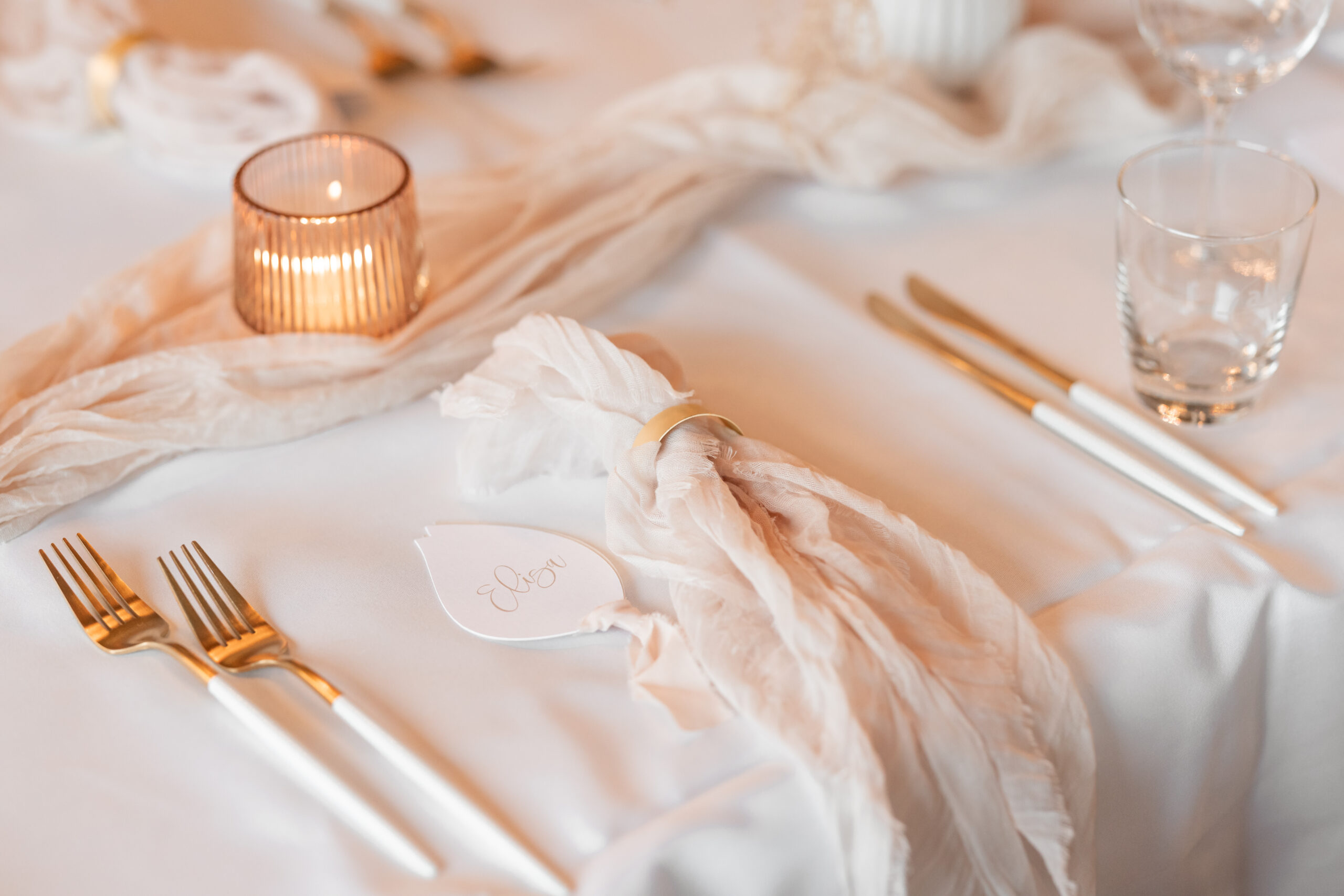 DekoandDesign_Hochzeit_Heuchelberger_Warte_Besteck_Servietten_Tischläufer_Musselin_leihen (2) - Kopie Ein durchsichtiger Glasteller mit einer gefalteten magentafarbenen Serviette und einer orangefarbenen Blume darauf steht neben einer orangefarbenen Speisekarte. Auf beiden Seiten des Tellers liegt goldfarbenes Besteck. Im Hintergrund sind Blumen zu sehen.