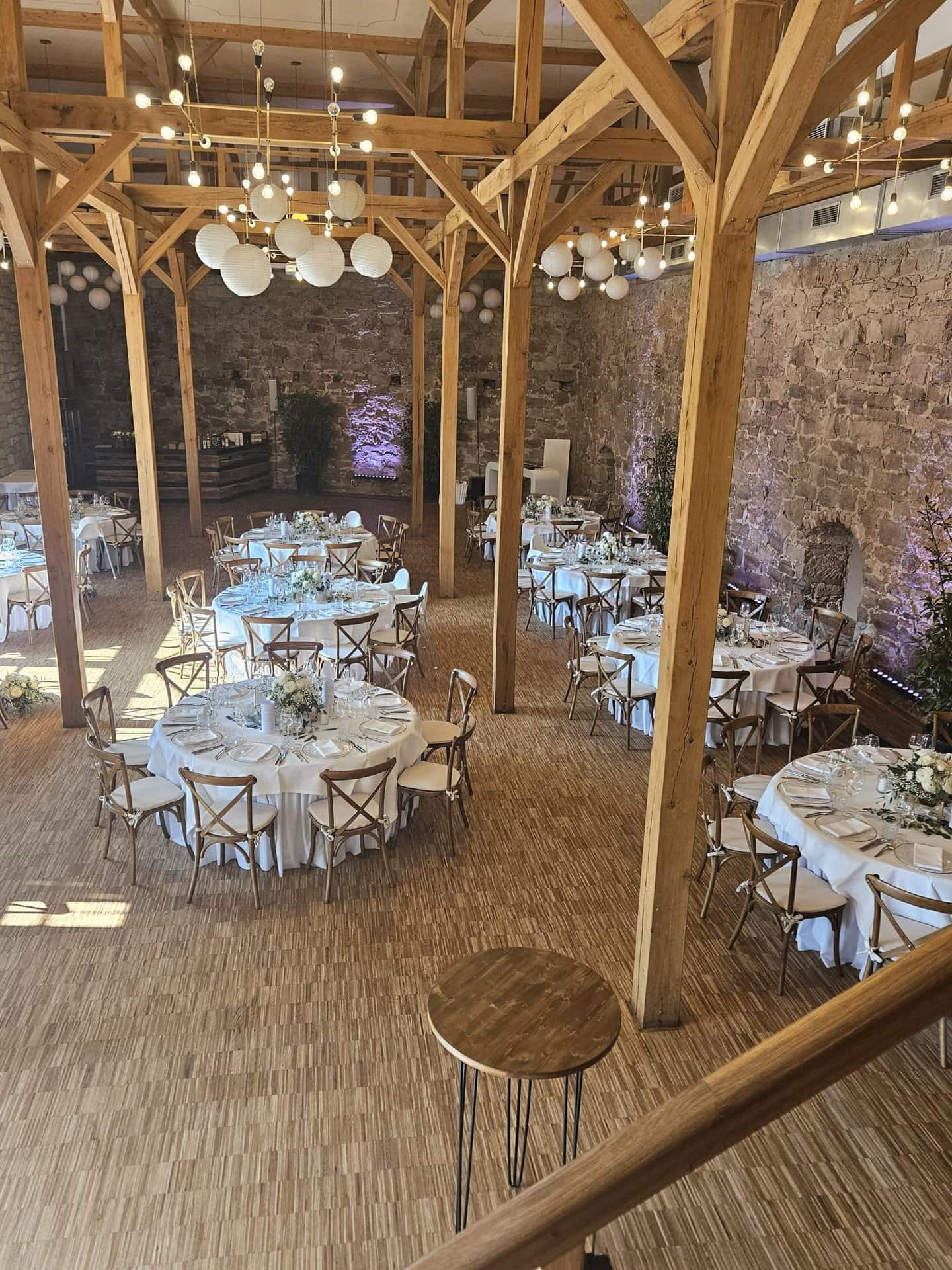 Hoher Veranstaltungssaal im Schloss Michelfeld mit Holzbalken, mehreren runden Esstischen mit weißen Tischdecken und von den Dachsparren hängenden Kugellampen. Steinmauern und Bäume schmücken den