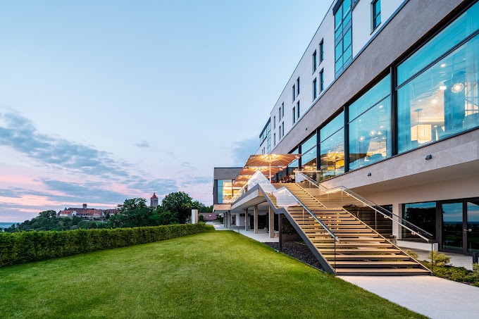 Außenansicht einer modernen Orangerie in der Abenddämmerung mit beleuchtetem Innenraum und Treppe vor einem Abendhimmel.