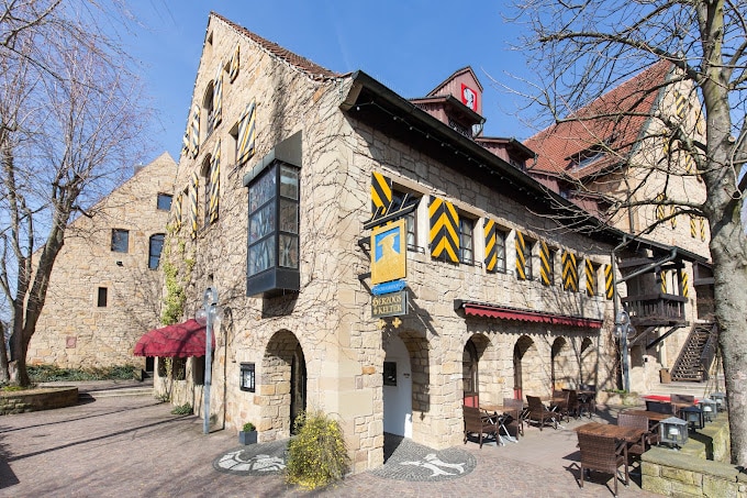 Altes Steingebäude mit Fachwerkanbau, in dem sich ein Restaurant befindet. Es gibt Tische draußen unter roten Sonnenschirmen, mit Bäumen in der Nähe an einem sonnigen Tag an der Herzogskelter Güglingen