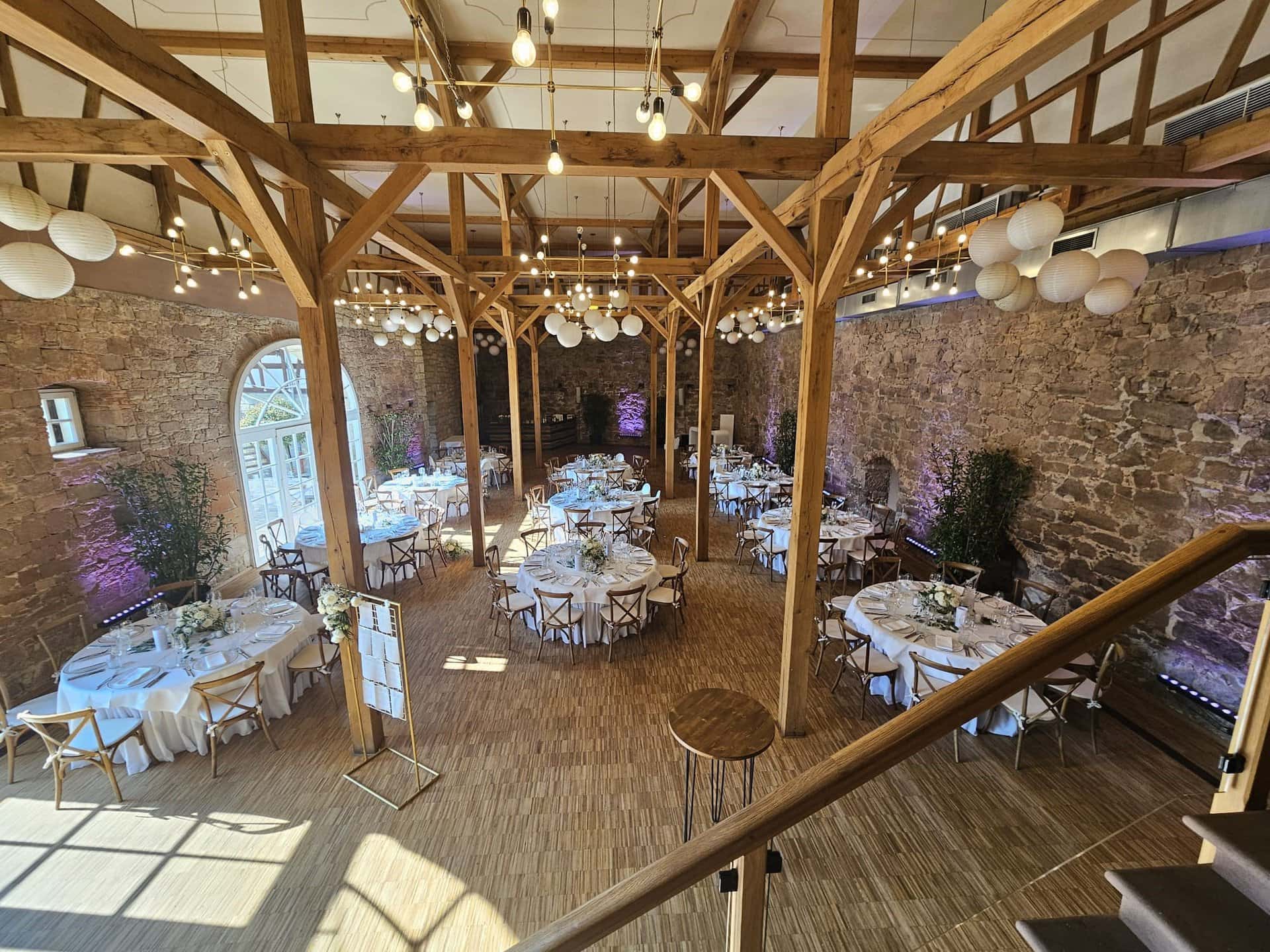 Innenansicht eines rustikalen Hochzeitslokals im Landgut Schloss Michelfeld mit Holzbalken, Steinwänden und runden Tischen für einen Empfang, beleuchtet mit Tageslicht und Hängelampen.