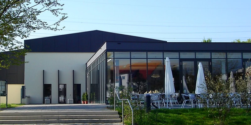 Modernes Gebäude mit großer Glasfassade und schattigem Sitzbereich mit Sonnenschirmen und Tischen in Rauers Guter Stube.