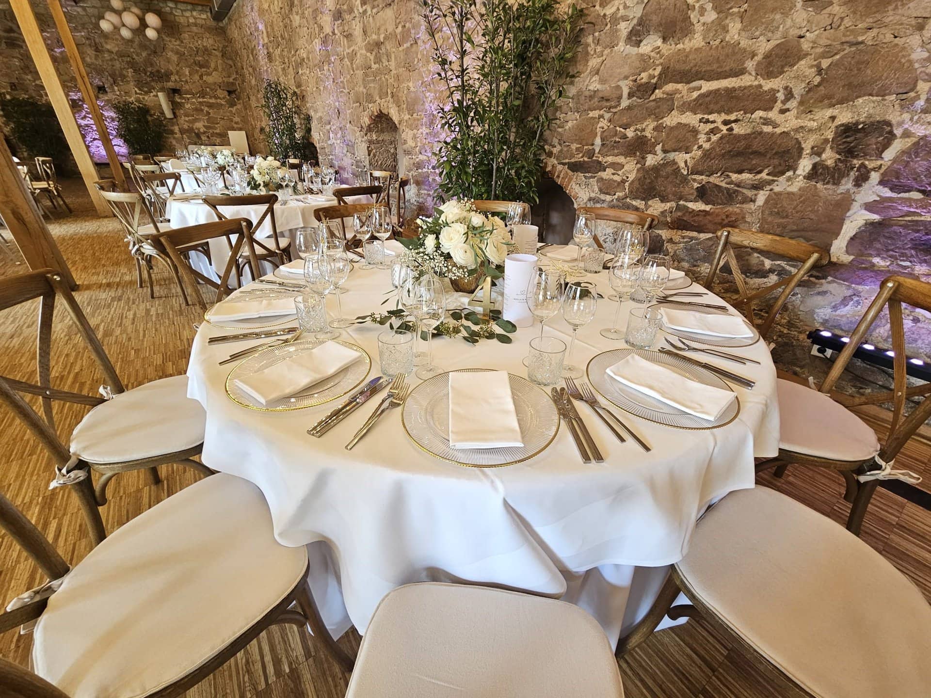 Elegantes Speisezimmer für eine Hochzeit im rustikalen Steinsaal des Landgut Schlosses, mit runden Tischen mit weißen Tischdecken, Blumenschmuck und formellem Essgeschirr.
