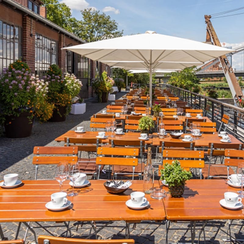 Essbereich im Freien mit Holztischen unter einem großen weißen Sonnenschirm an einem sonnigen Tag, perfekt für malerische Orte.
