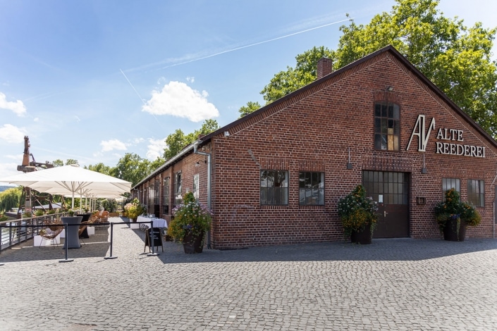 Backsteingebäude mit Schild „Reblandhalle“ und Außensitzplatz unter weißem Sonnenschirm.