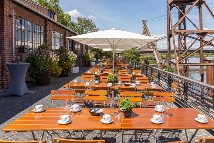 Gastgarten mit gedeckten Holztischen unter einem großen Sonnenschirm, mit Blick auf den Fluss in der Nähe der Alten Reederei Heilbronn und Industriebauten.