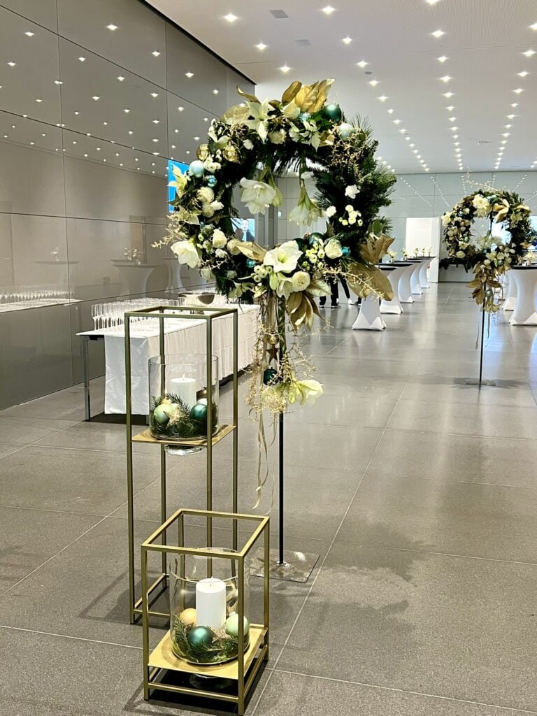 Elegante Feiertagsdekoration bestehend aus einem großen Kranz, geschmückt mit weißen Blumen und grünen Blättern auf einem Ständer, begleitet von einer Kerzenpräsentation in einer modernen Innenumgebung.