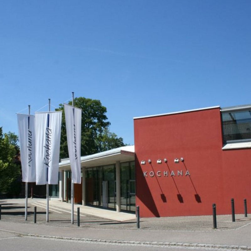 Ein modernes Gebäude mit roter Fassade und im Wind flatternden weißen Bannern mit der Aufschrift „Kochana“ dient im Gebietsführer als Orientierungspunkt.