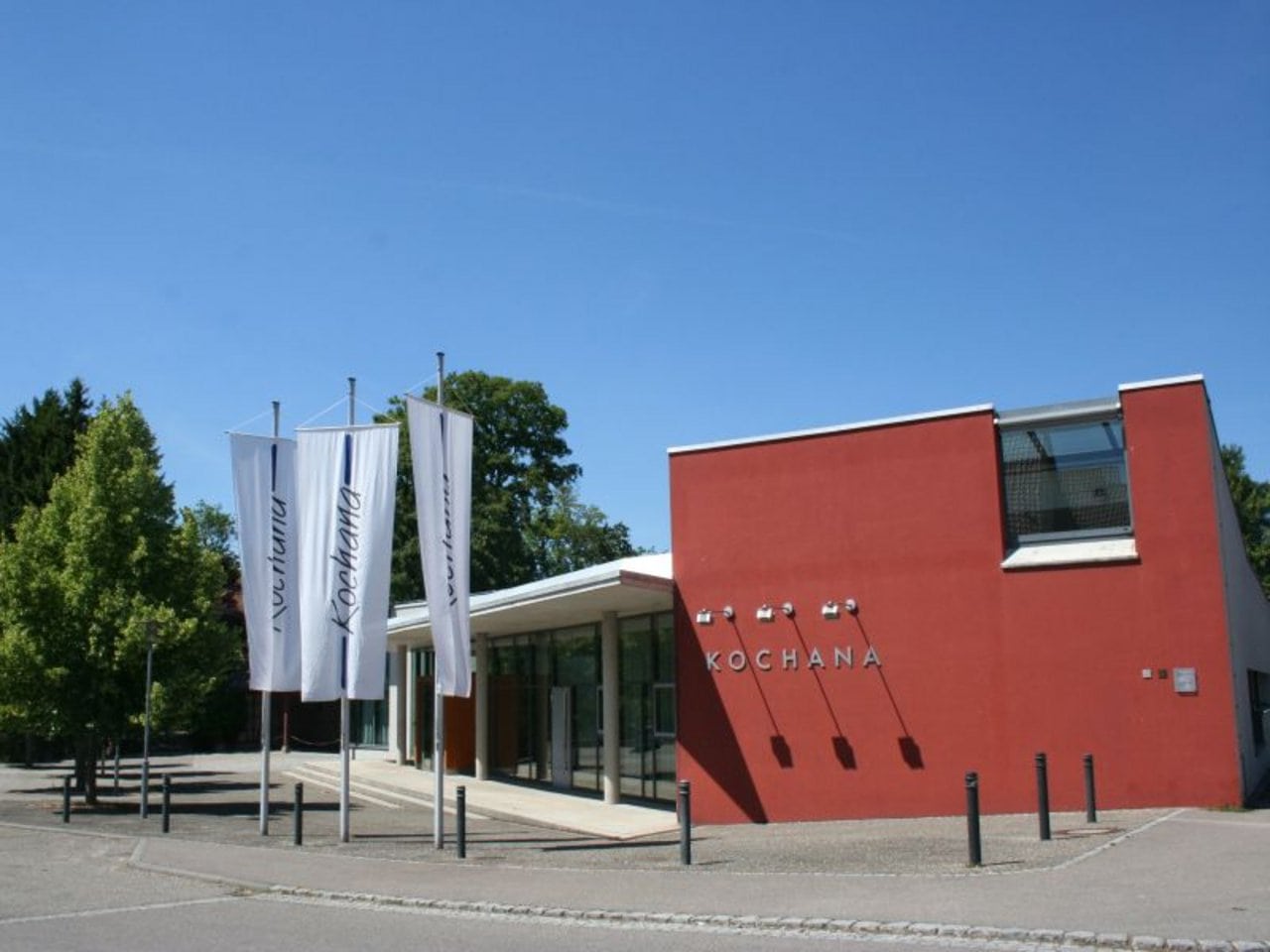 Modernes Festhallengebäude mit Flaggen und der Aufschrift „kochana“ an der Fassade unter einem strahlend blauen Himmel.