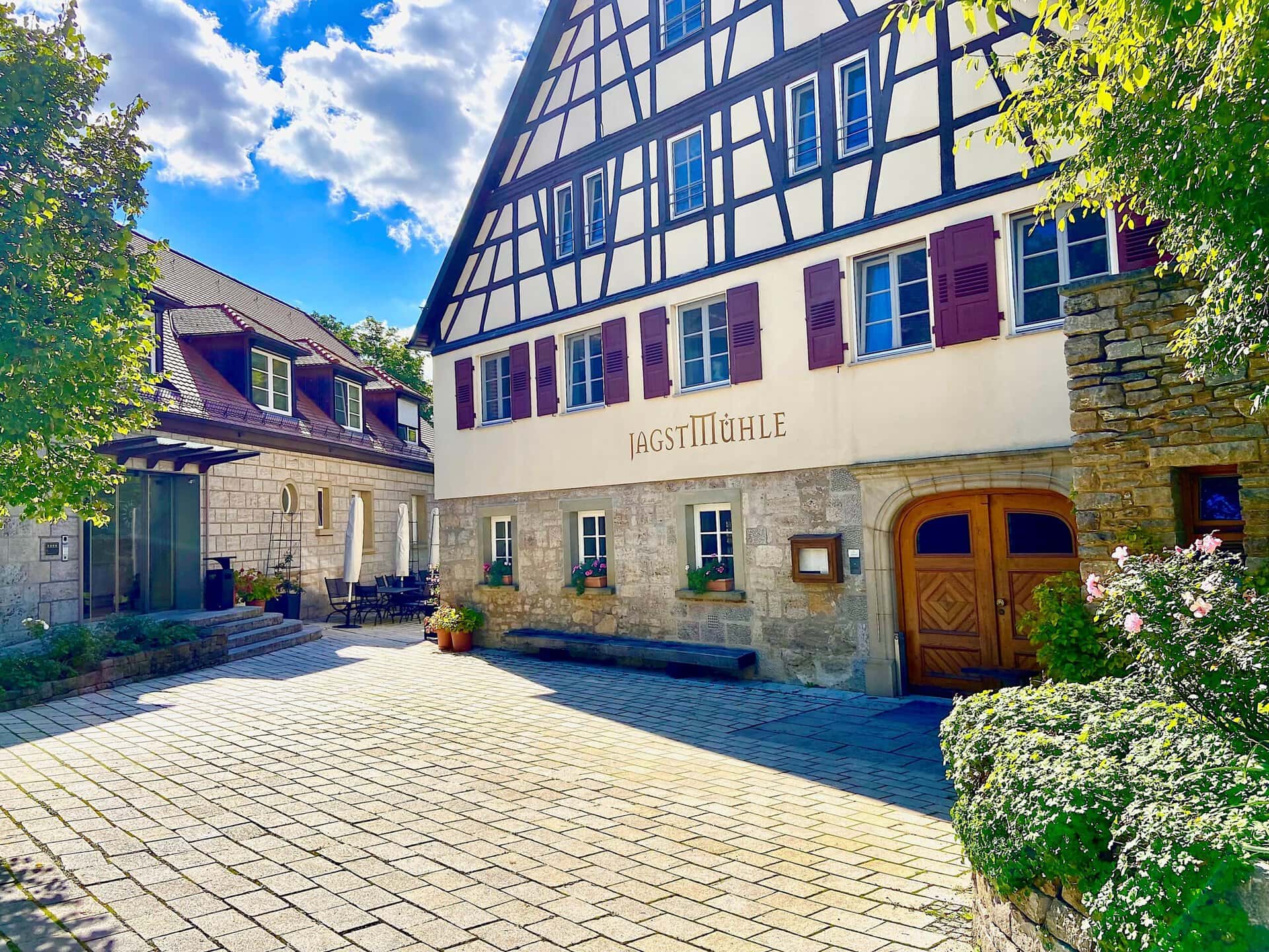 Traditionelles Fachwerkhaus mit der Aufschrift „Landgasthof Jagstmühle“ neben einem Steingebäude unter einem strahlend blauen Himmel.