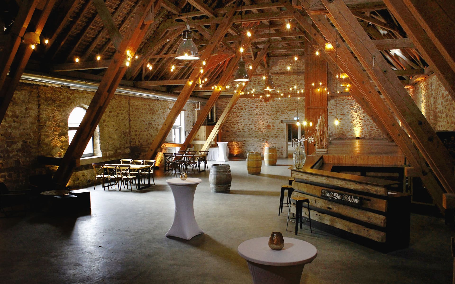 Rustikales Scheunen-Interieur, das in einen Veranstaltungsraum mit freiliegenden Holzbalken und Backsteinwänden umgewandelt wurde, mit stimmungsvoller Beleuchtung und einer Mischung aus modernen und Vintage-Möbeln. Willkommen in Ladenburgs Eventlocation Eichenst