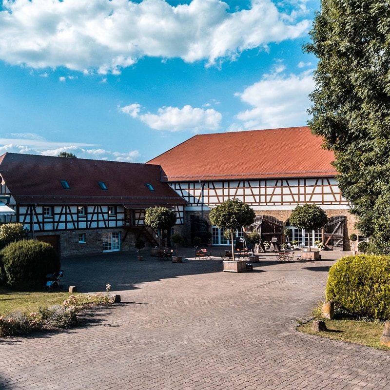 Traditionelles Fachwerkgebäude mit rotem Dach, an malerischen Orten gelegen, umgeben von viel Grün unter einem blauen Himmel mit Wolken.
