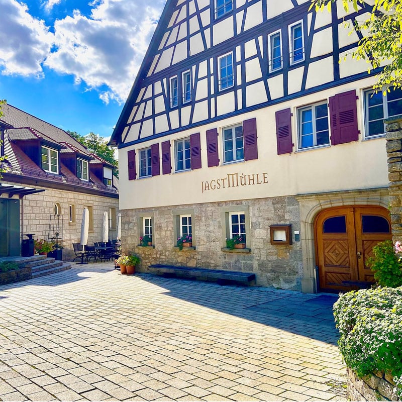 Traditionelles Fachwerkhaus in malerischer Lage mit dem Schild „Gaststätte Hühle“ über der Eingangstür, Kopfsteinpflaster und blauem Himmel.