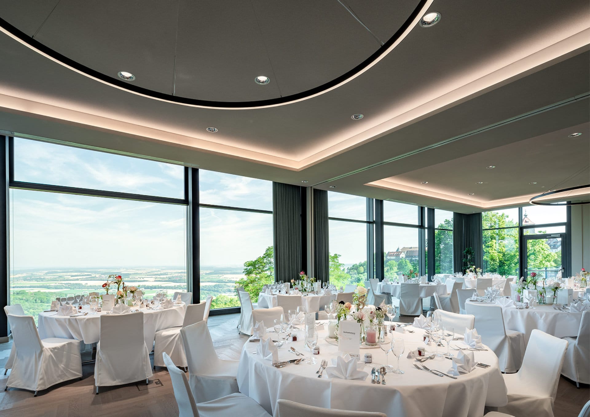 Ein eleganter Bankettsaal im Panoramahotel mit runden, für eine Veranstaltung gedeckten Tischen und großen Fenstern, die einen Panoramablick auf die Waldenburger Landschaft draußen bieten.