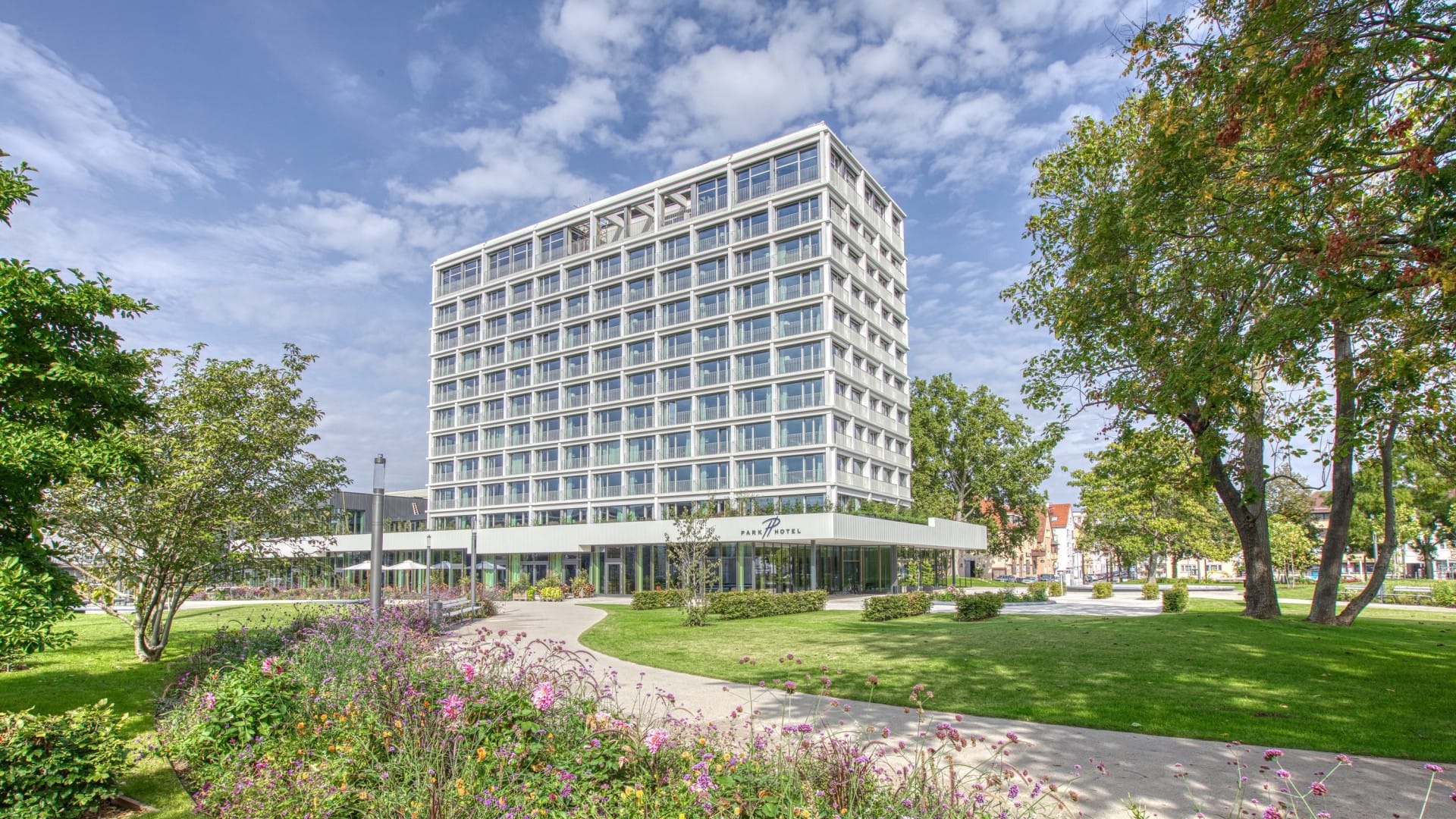 Modernes Bürogebäude mit Blick auf einen angelegten Stadtpark mit üppigem Grün und Fußwegen unter strahlend blauem Himmel, in der Nähe der Luxusunterkünfte des Parkhotels Heilbronn.