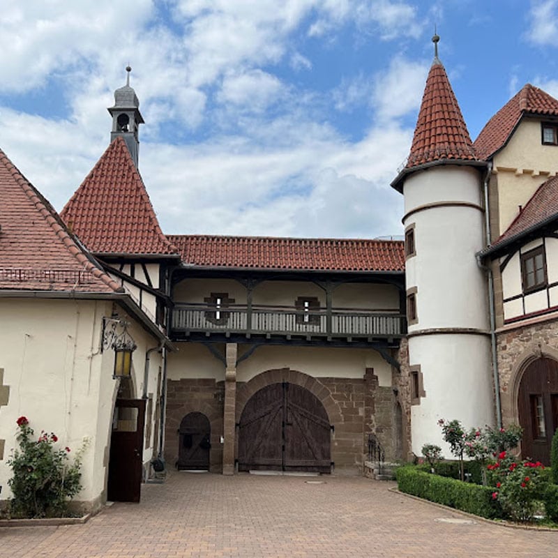Traditionelle europäische Orte mit einem Gebäude mit einem Holzbalkon, turmartigen Strukturen und einem gewölbten Eingang unter einem teilweise bewölkten Himmel.