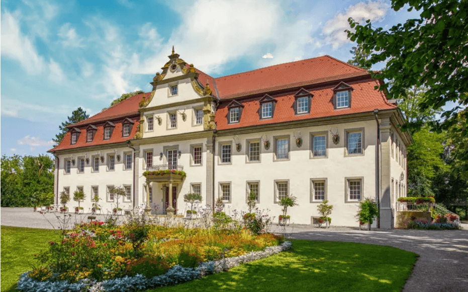 Ein Herrenhaus im Barockstil, umgeben von gepflegten Gärten unter klarem Himmel, das heute als Hotel Wald- und Schlosshotel Friedrichsruhe dient.