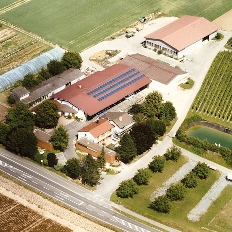 Luftaufnahme eines ländlichen Bauernhofs mit landwirtschaftlichen Gebäuden, Solarmodulen und angrenzenden Feldern an verschiedenen Standorten.