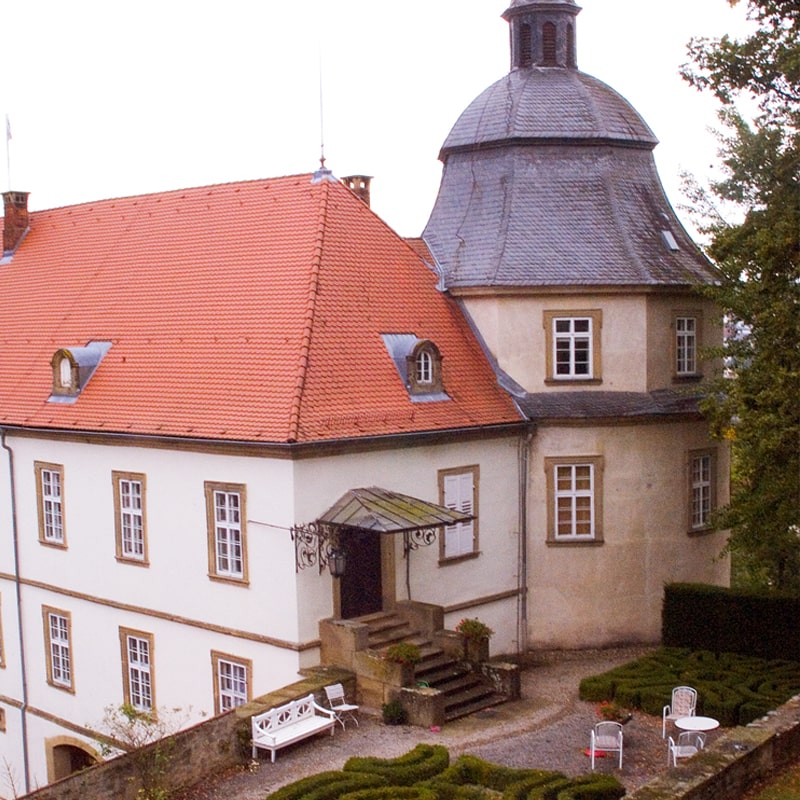 Luftaufnahme eines historischen Gebäudes an mehreren Standorten mit einem roten Ziegeldach und einem kleinen Garten mit gepflegten Büschen.