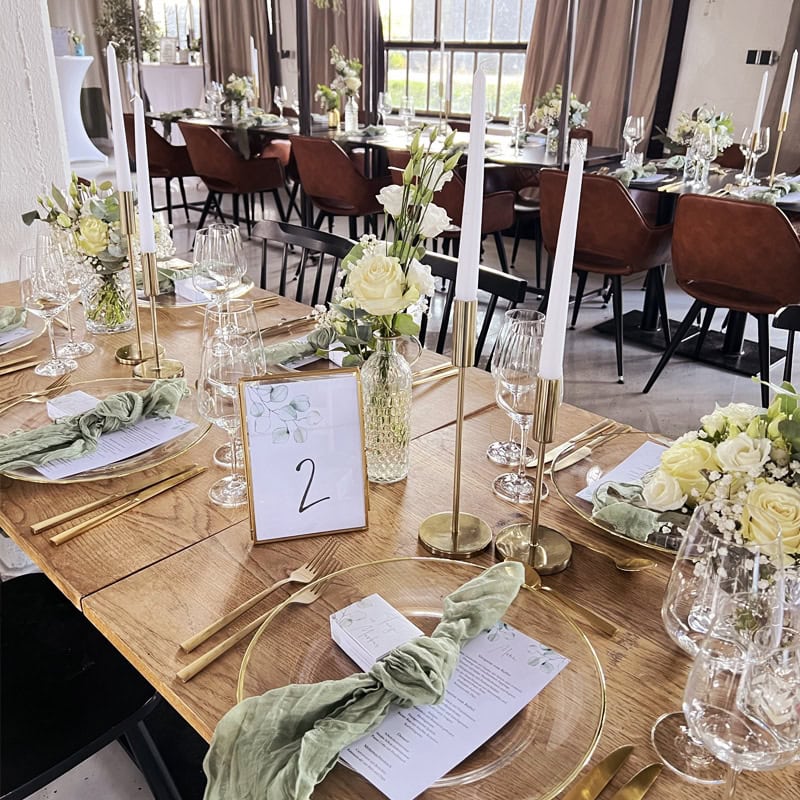 Elegante Tischdekoration für den Hochzeitsempfang mit floralen Tischdekorationen, goldenem Besteck und nummerierten Karten in einem stilvollen Veranstaltungsort.