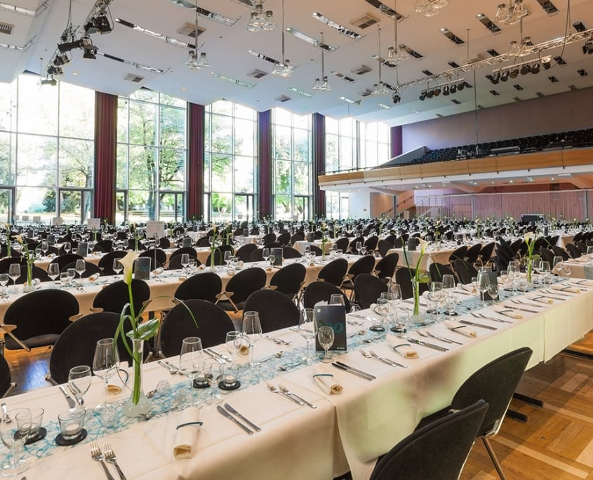 Ein großer Bankettsaal in der Harmonie Heilbronn, ausgestattet mit zahlreichen Esstischen, jeder mit weißem Tuch bedeckt, mit schwarzen Stühlen ausgestattet und mit blauen Mittelstücken dekoriert.