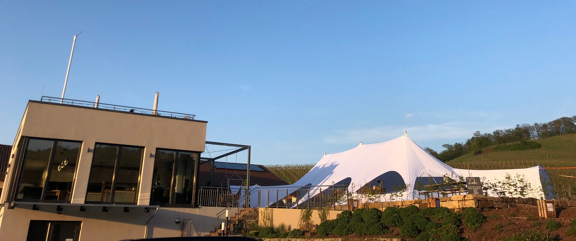 Modernes Gebäude mit Dachterrasse und einem großen weißen Zelt, das bei Sonnenuntergang auf einer angrenzenden Wiese mit Blick auf die weitläufigen Weinberge des Weinguts Bihlmayer aufgebaut wird.