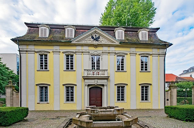 Zweistöckiges gelb-weißes Gebäude im Barockstil mit zentralem Eingang und symmetrischen Fenstern, flankiert von Bäumen und einem kleinen Brunnen davor, bekannt als Heilbronner Schießhaus