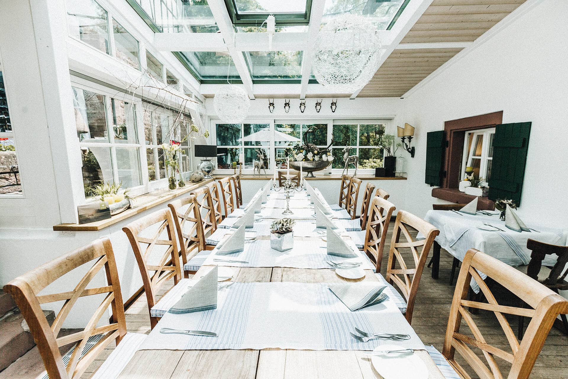Heller und luftiger Speiseraum im Landgasthof Heidersbacher Mühle mit einer langen, gedeckten Tafel, umgeben von Holzstühlen, unter einem Glasdach.