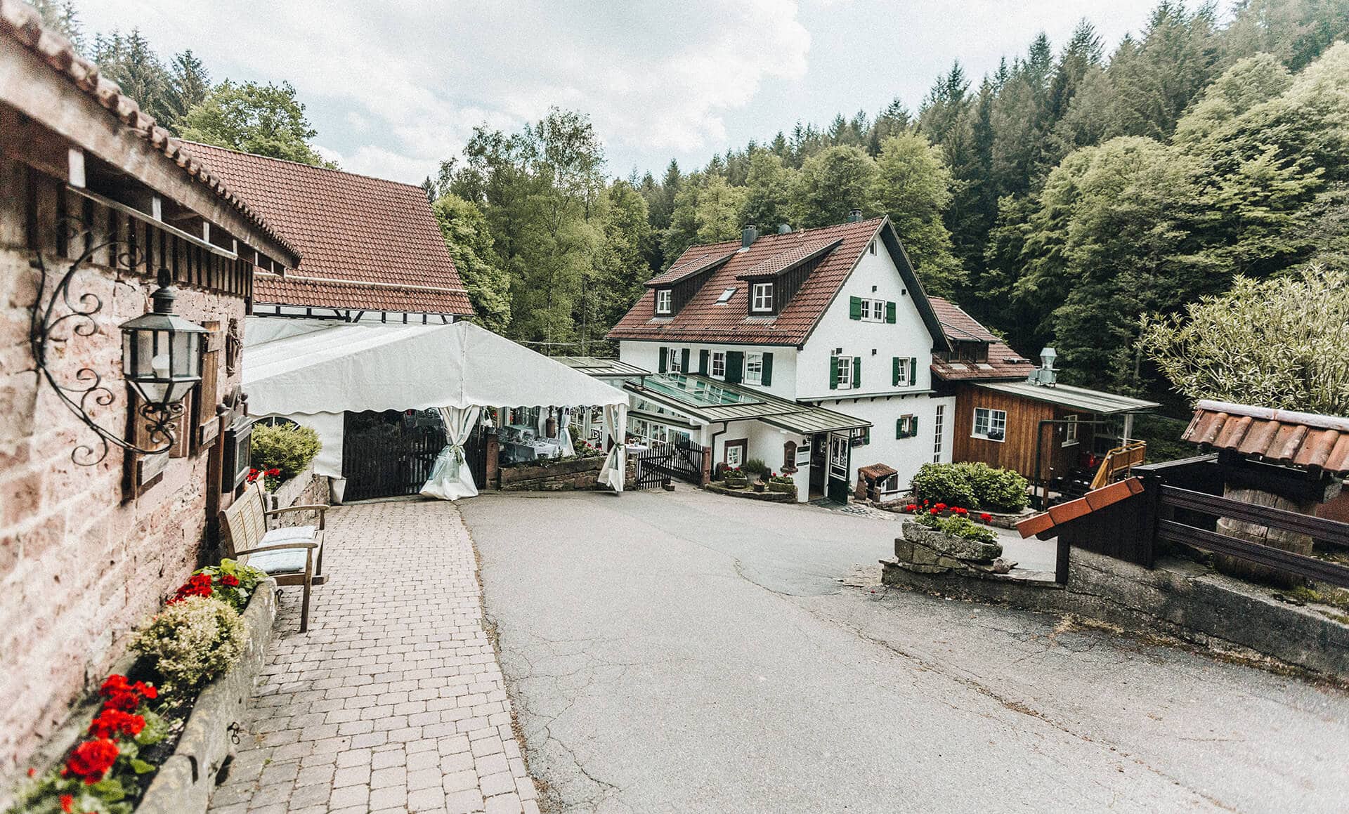 Eine malerische Dorfszene mit einem traditionellen weißen Haus, heute der Landgasthof Heidersbacher Mühle, umgeben von üppigem Grün und einer Kopfsteinpflasterstraße.