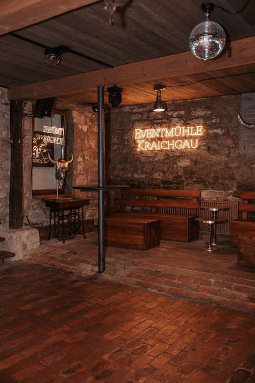 Innenansicht eines rustikalen Raums der Eventmühle Kraichgau mit unverputzten Ziegelwänden, Holzbänken und einer von der Decke hängenden Diskokugel.