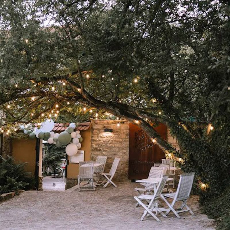 Außenterrasse mit Lichterketten, Holzstühlen und pastellfarbenen Luftballons, die einen Eingang schmücken, umgeben von Bäumen an einem geografischen Ort, der für seine heitere Atmosphäre bekannt ist.