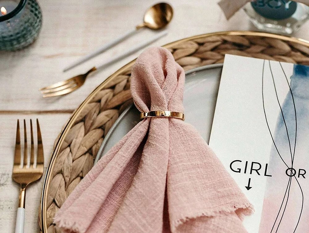 Tischdekoration mit einer rosa Serviette, die von einem goldenen Ring auf einem weißen Teller gehalten wird, begleitet von einer goldfarbenen Gabel und einem Löffel und einer Karte mit der Aufschrift „GIRL OR“ mit dekorativen Elementen. Ideal für diejenigen, die Tischdekoration mieten möchten.