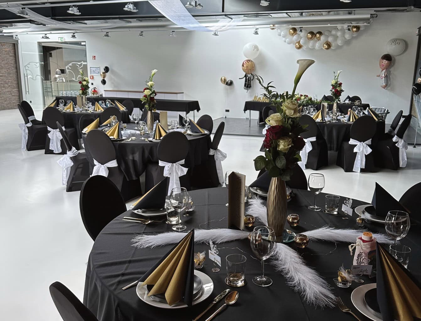 Ein dekorierter Veranstaltungsort mit schwarzen, runden Tischen mit goldenen Spitzhüten, schwarzen Stühlen mit weißen Schärpen, floralen Tafelaufsätzen und hängenden Dekorationen in Gold, Weiß und Schwarz – perfekt für Feiern oder Firmenveranstaltungen von SML Carlocation.
