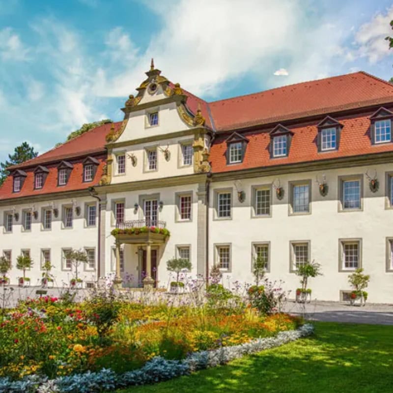 Ein symmetrisches Gebäude mit rotem Ziegeldach und weißer Fassade steht stolz zwischen verschiedenen Veranstaltungsorten, umgeben von einem gepflegten Garten voller bunter Blumen und Grünpflanzen. Der Himmel darüber ist klar mit ein paar Wolken, was die Schönheit dieser malerischen Orte noch verstärkt.